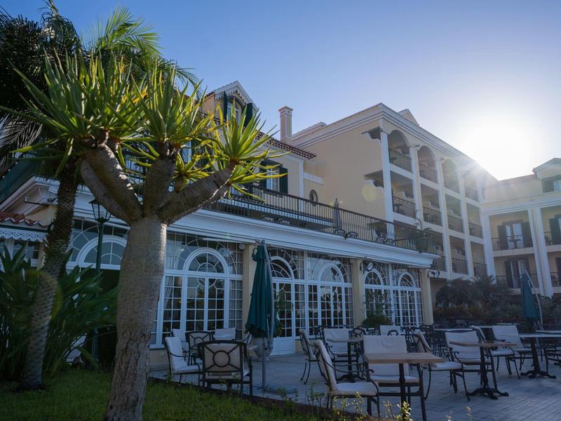 Mehrstöckiges Hotel mit großen Fenstern, Außenbestuhlung und Palmen am frühen Abend.