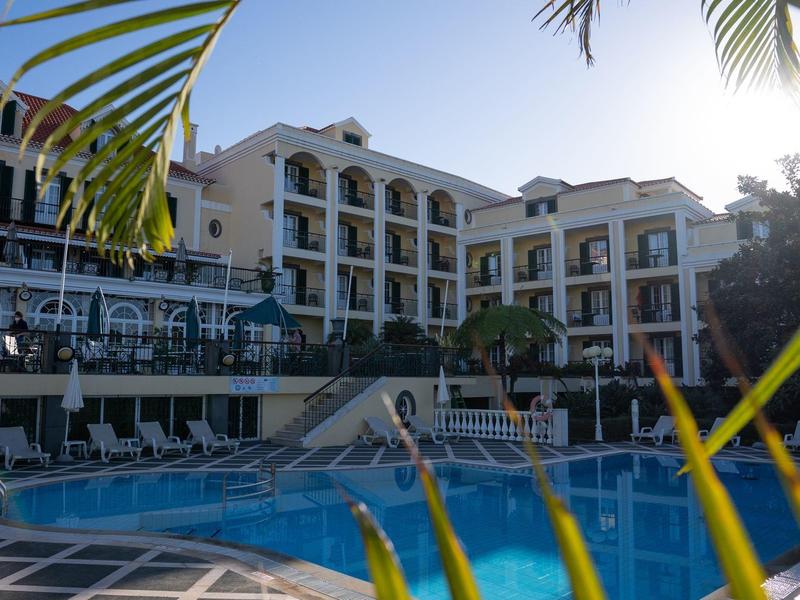 Großer Außenpool vor mehrstöckigem Hotelgebäude mit Balkon, umgeben von Palmen und Sonnenliegen.