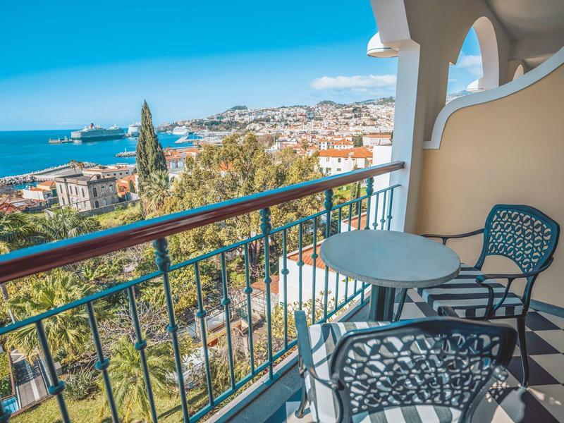 Balkon mit zwei Stühlen und Tisch mit Blick auf Stadt und Meer bei klarem Himmel.