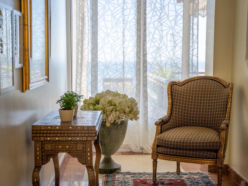 Gemütliche Ecke mit gepolstertem Sessel, hölzernem Beistelltisch, Blumenvasen und hellen Spitzenvorhängen.