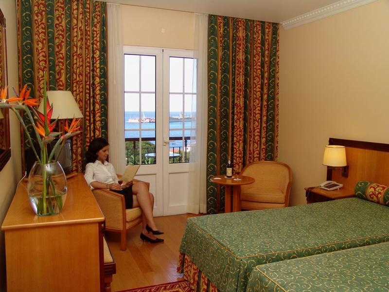 Hotelzimmer mit Doppelbett, grünem Bettbezug, Sitzbereich, Tisch, Blumen und Blick auf das Meer.