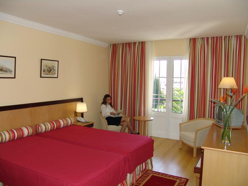 Ein Zimmer mit roten Betten, einer Frau am Fenster, Holzboden und gestreiften Vorhängen.