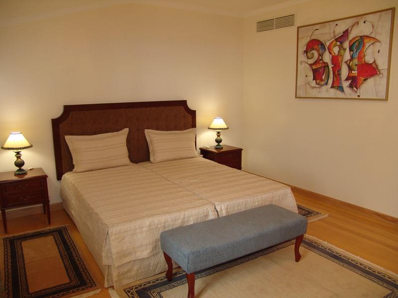 Ein schlichtes Schlafzimmer mit Doppelbett, zwei Nachttischen mit Lampen, Hocker und moderner Wandkunst.