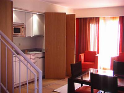 Pequeña cocina de hotel con área de comedor, armarios blancos y sillón rojo junto a ventana con cortinas.