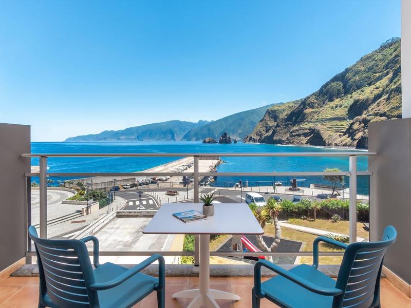 Balkon mit zwei Stühlen und Tisch, Blick auf Meer, Küste und Berge bei klarem Himmel.