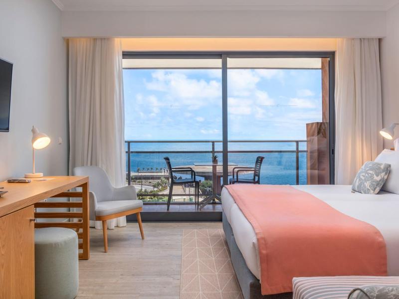 Helles Hotelzimmer mit Doppelbett, Terrasse und Meerblick bei Tag.