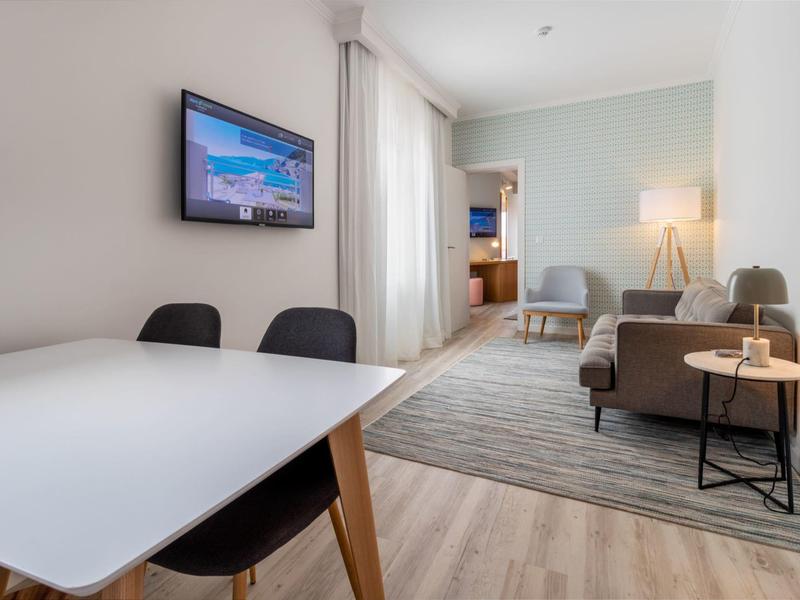 Moderner Wohnraum mit weißem Tisch, schwarzen Stühlen, grauem Sofa, Teppich und Wand-TV.