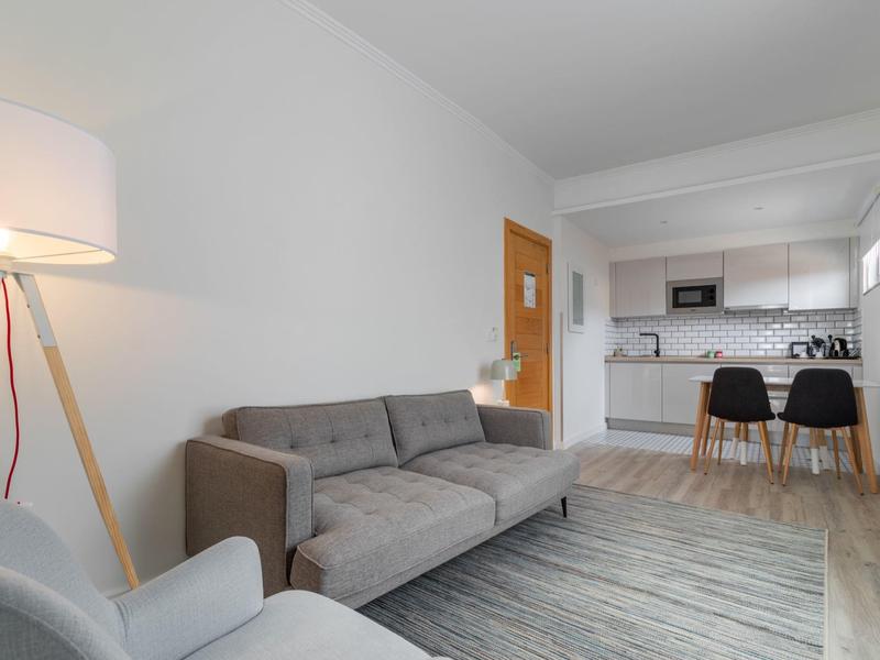 Moderner Wohnraum mit grauem Sofa, hellem Teppich, Stehlampe und offener Küche mit Essbereich.