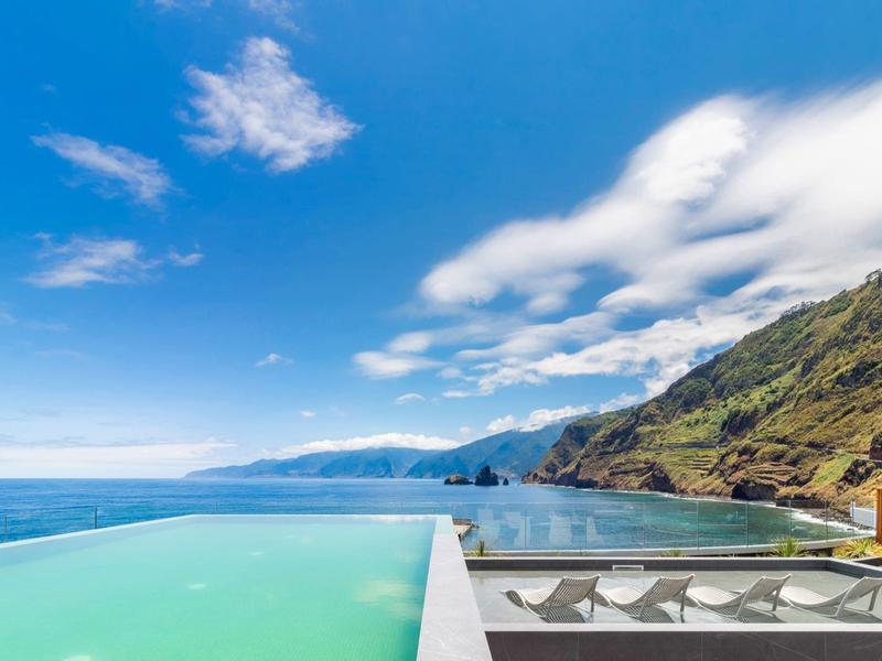 Infinity-Pool mit türkisblauem Wasser vor Bergen, Meer und blauem Himmel mit weißen Wolken.