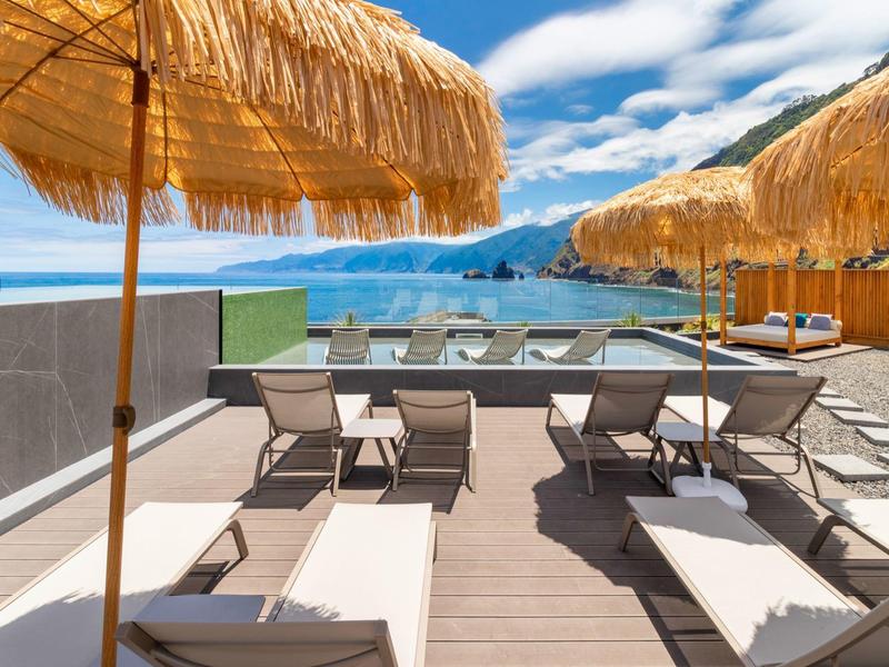 Dachterrasse mit Liegestühlen, Strohhächern und Blick auf blauen Himmel und Meer.