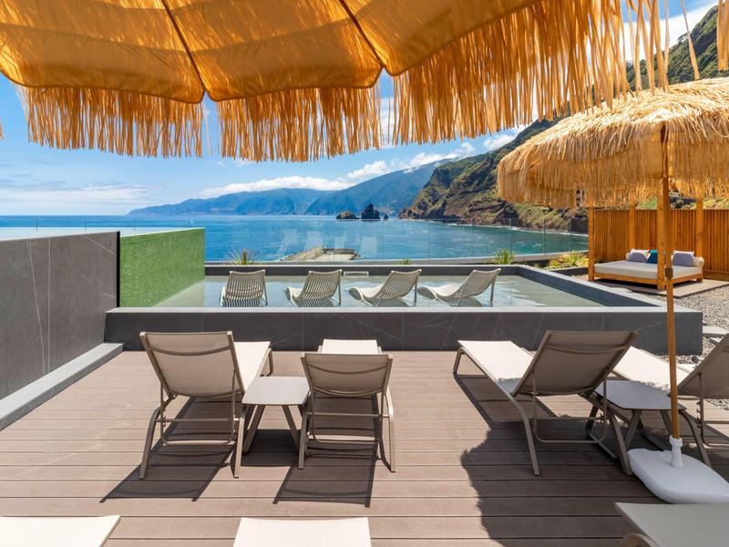 Terrasse mit Sonnenliegen, Tischen und Strohmusterschirmen mit Blick auf Meer und Berge.