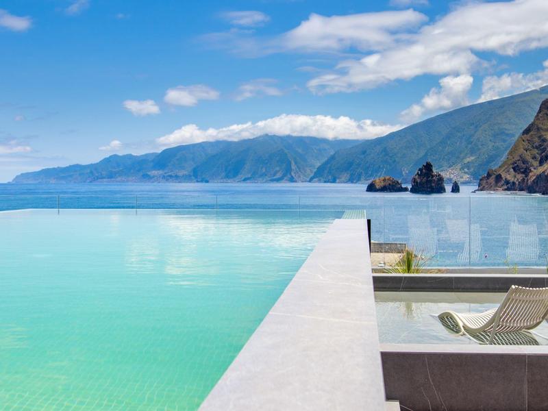 Endloser Pool mit türkisfarbenem Wasser, zwei Liegestühlen und Bergen im Hintergrund.