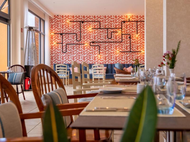 Modernes Restaurant mit hellen Holzmöbeln, dekorativer Wandbeleuchtung und großen Fenstern.