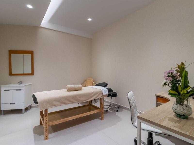 Heller Behandlungsraum mit Massageliege, weißem Stuhl, Kommode und Blumendekoration.