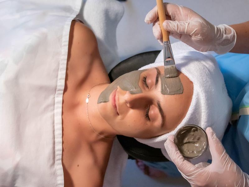 Eine Frau liegt entspannt, trägt ein Handtuch auf dem Kopf, während ihr Gesicht mit einer Maske bestrichen wird.