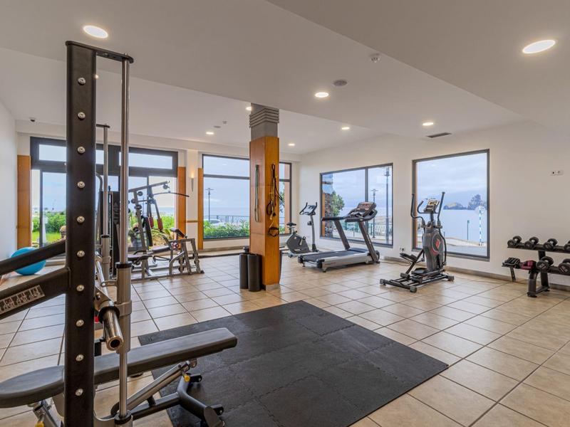 Moderner Fitnessraum mit Geräten, großen Fenstern, heller Beleuchtung und Kachelboden.