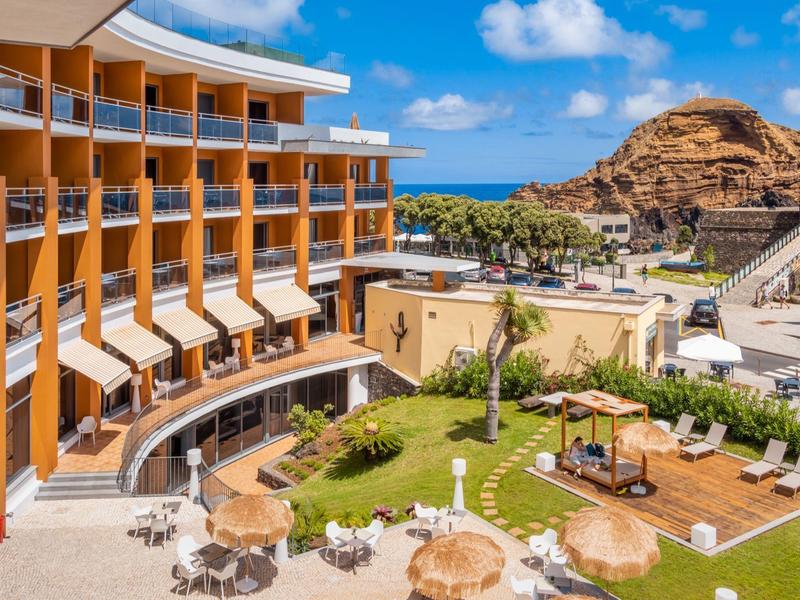 Hotel mit geschwungener Fassade, Außensitzbereich, Liegestühlen und Blick auf Hügel und Meer.