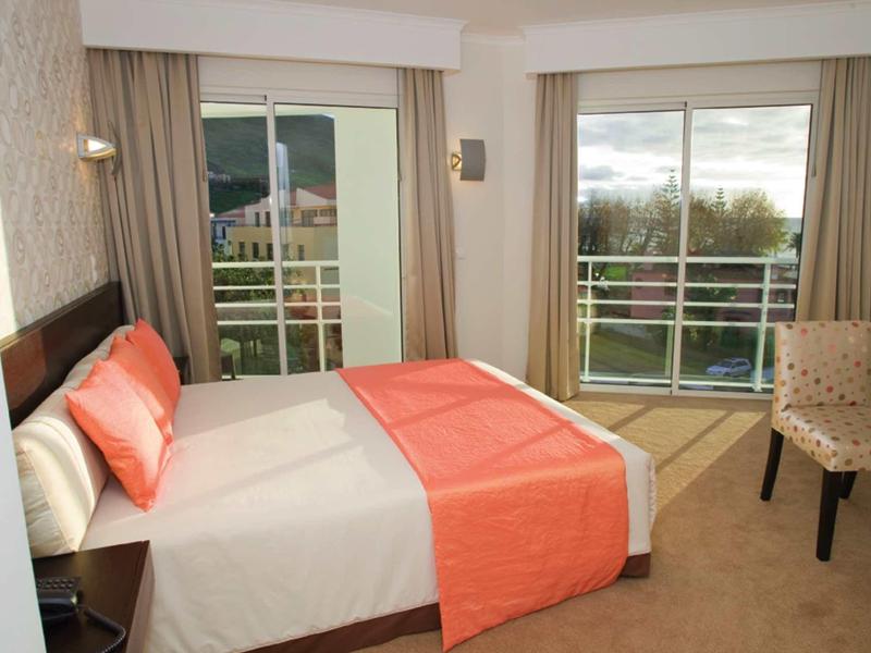 Camera d'hotel con letto matrimoniale, biancheria da letto arancione e due grandi finestre con vista su una terrazza.