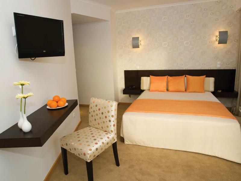 Piccola camera d'hotel con letto matrimoniale, cuscini arancioni e colori tenui.