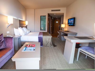 Modernes Hotelzimmer mit Doppelbett, Schreibtisch, Couch und Fernseher, heller Raum in sanften Farben.