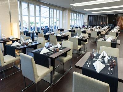 Modernes Restaurant mit hellen Sesseln, dunkel gedeckten Tischen und großem Fensterfrontbereich.