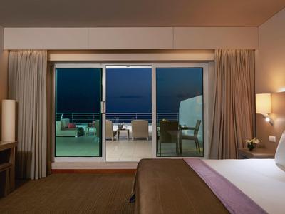 Modernes Hotelzimmer mit Doppelbett, Balkon und Meerblick bei Nacht, warme Beleuchtung innen.