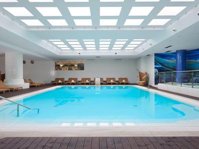 Großes, rechteckiges Hallenbad mit hellem, blau leuchtendem Wasser und bequemen Liegestühlen.