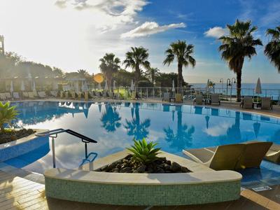 Hotelpool mit Palmen, Sonnenliegen und Meerblick bei Sonnenschein am Nachmittag.