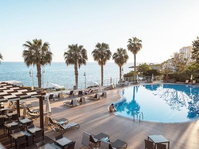 Poolbereich mit Liegestühlen, Sonnenschirmen, Palmen und Meerblick an sonnigem Tag.