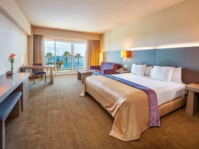 Helles Hotelzimmer mit großem Bett, Schreibtisch, Sessel und Balkon mit Blick auf Palmen.