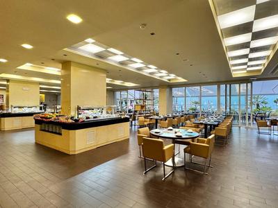 Modern hotel restaurant with buffet stations, large windows, and tables with chairs.