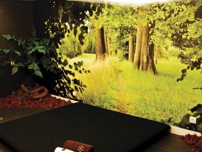 Ein Raum mit schwarzem Bett, dekorativem Laub und einer Wand mit großem Waldgemälde.