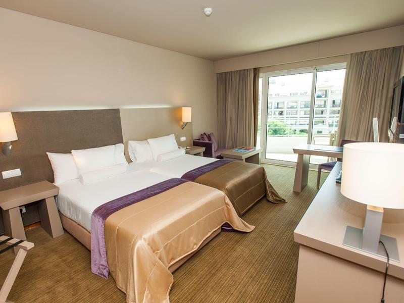 Helles Hotelzimmer mit Doppelbett, zwei Nachttischen, Schreibtisch, Stuhl und großem Fenster mit Vorhängen.