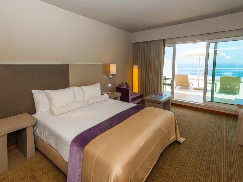 Hell eingerichtetes Hotelzimmer mit großem Bett, Schreibtisch, Sesseln und Meerblick vom Balkon.