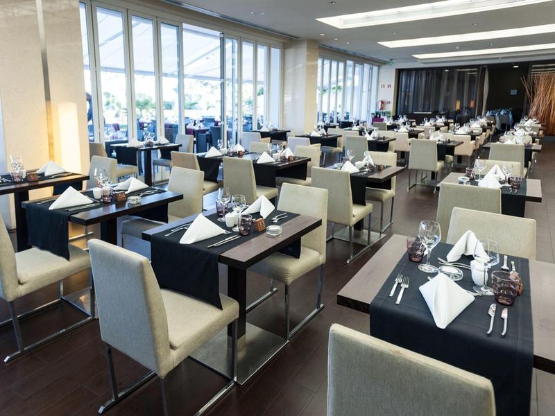 Modernes Restaurant mit hellen Sesseln, dunkel gedeckten Tischen und großem Fensterfrontbereich.