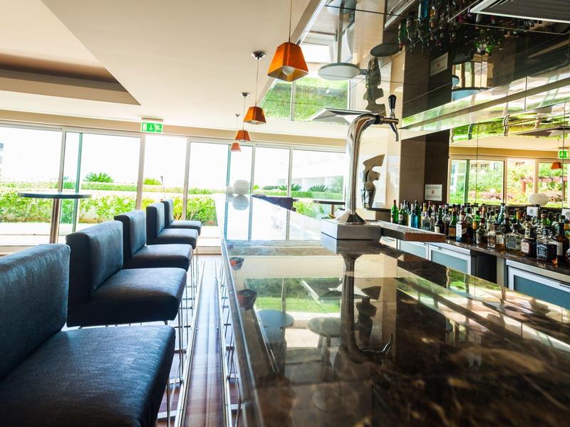 Moderner Barbereich mit glänzendem Holztheken, blauen Sitzbänken und großen Fenstern.