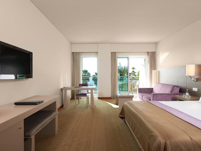 Modernes Hotelzimmer mit großem Bett, lila Sofa, Schreibtisch, Fernseher und Balkon mit Pflanzen.