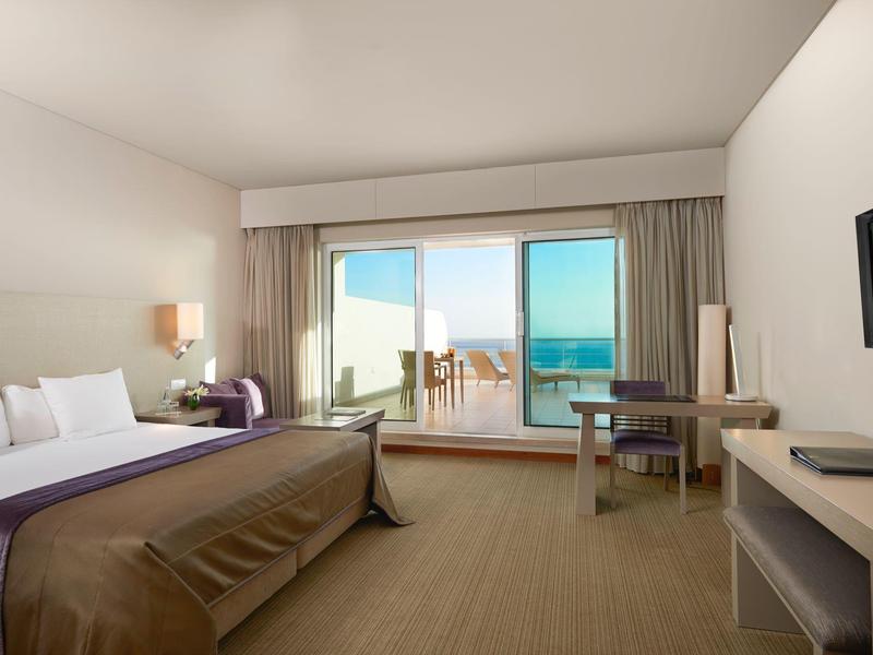 Helles Hotelzimmer mit Doppelbett, Schreibtisch, TV und Balkon mit Meerblick bei Tageslicht.