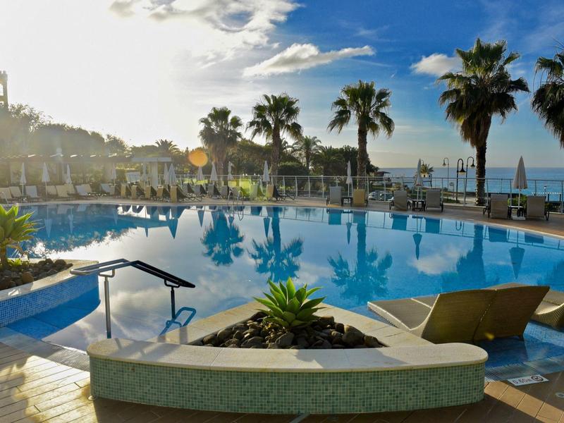 Hotelpool mit Palmen, Sonnenliegen und Meerblick bei Sonnenschein am Nachmittag.