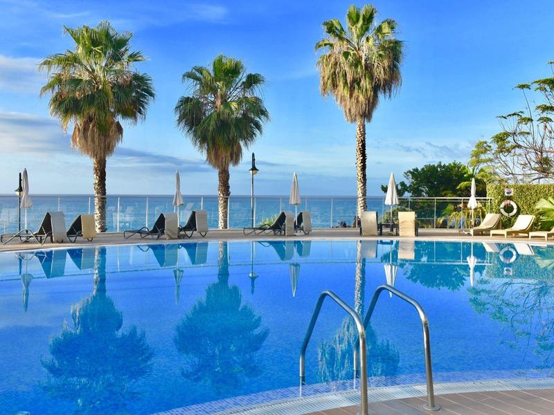 Schwimmbecken mit klar blauem Wasser, Palmen im Hintergrund und Sonnenschirmen am Rand.