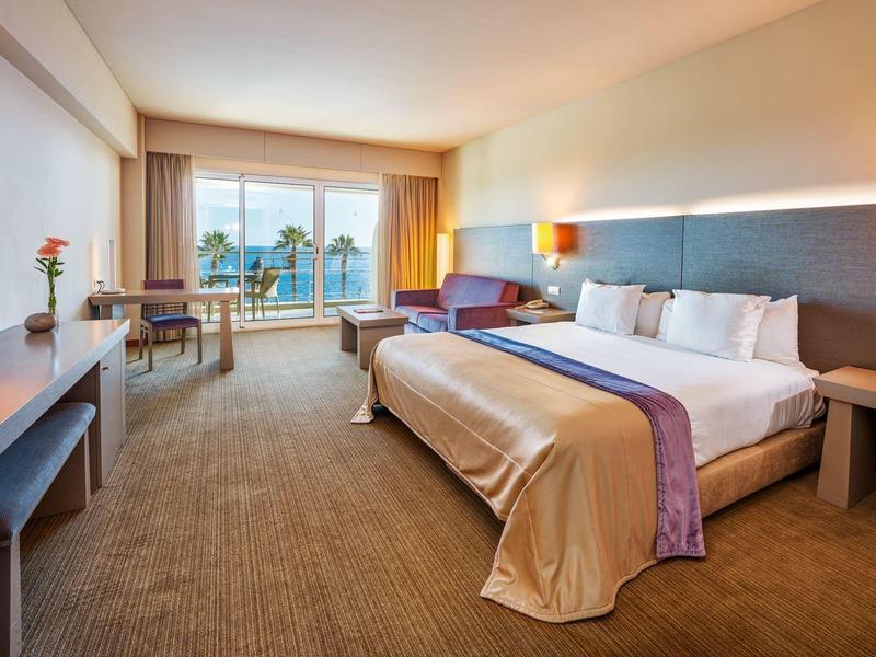 Helles Hotelzimmer mit großem Bett, Schreibtisch, Sessel und Balkon mit Blick auf Palmen.