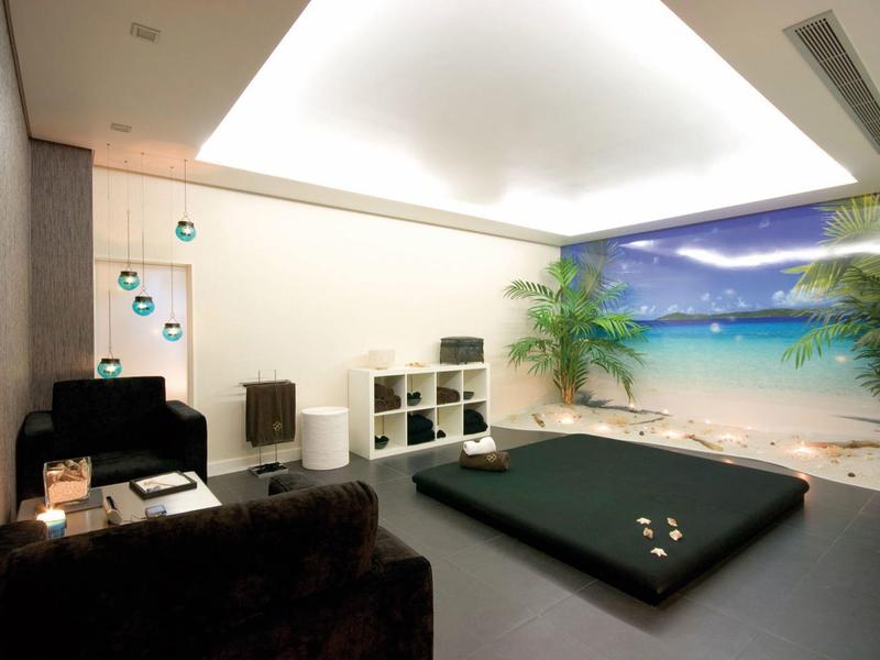 Gemütlicher Raum mit dunklem Sitz, flachem Bett und Wandbild von Strand mit Palmen.