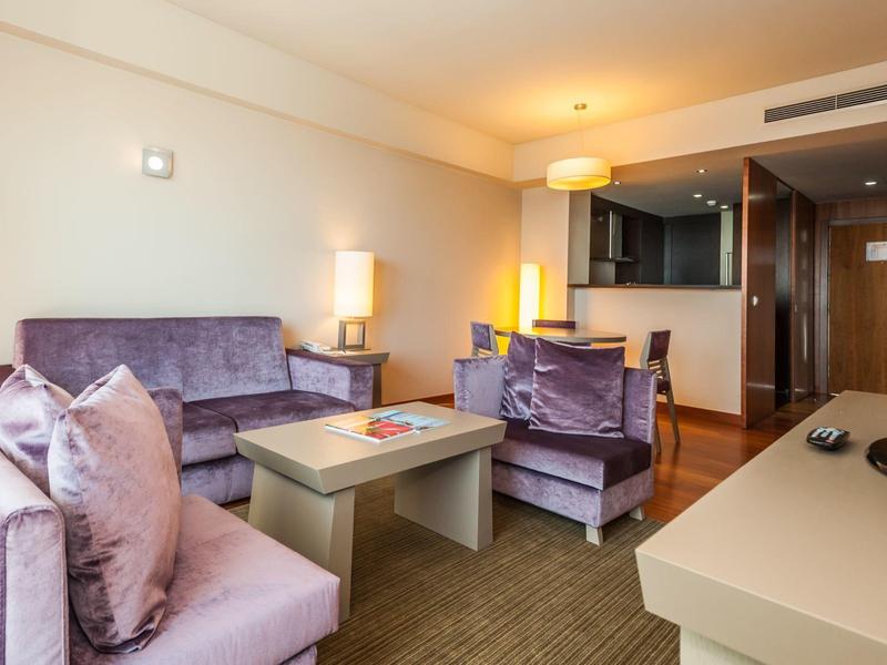 Gemütliches Wohnzimmer mit lila Sofa, Sesseln, Couchtisch und modernem TV-Bereich in warmem Licht.