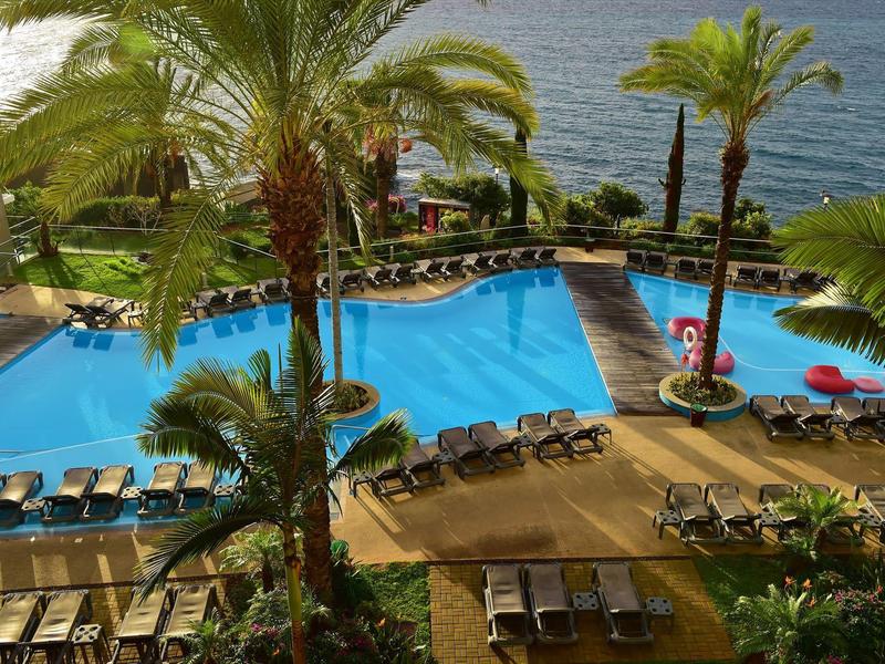 Großer Poolbereich mit Liegestühlen und Palmen neben dem Meer in einem Hotelresort.