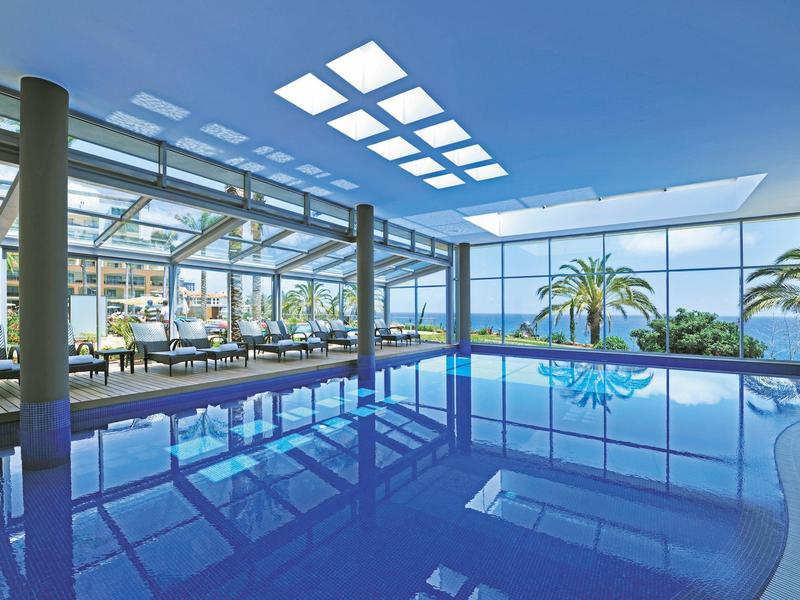 Innenpool mit Glasdach und Blick auf Palmen und das Meer bei klarem Himmel.