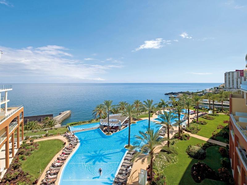 Blick auf einen Hotelpool mit Palmen und Meer im Hintergrund bei klarem Himmel.