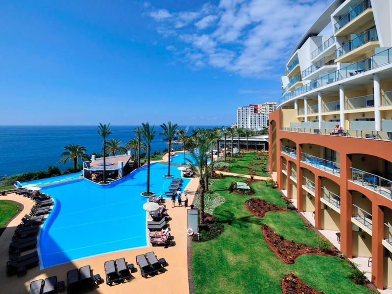 Hotelanlage mit großem Pool, Liegestühlen, Palmen und Meerblick bei sonnigem Himmel