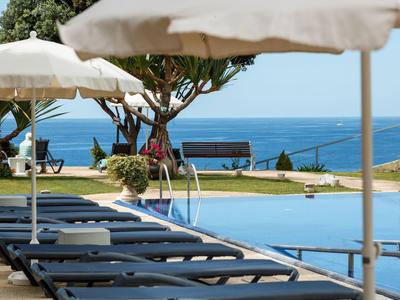 Leere Liegen mit weißen Sonnenschirmen neben einem Pool, im Hintergrund Meer und Bäume.