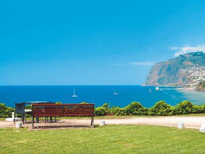 Bank mit Blick auf das blaue Meer, grünen Rasen und eine hohe Felsklippe im Hintergrund.
