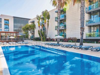 Moderner Außenpool mit blauer Wasserfläche, Liegestühlen und hellem Hotelgebäude im Hintergrund.